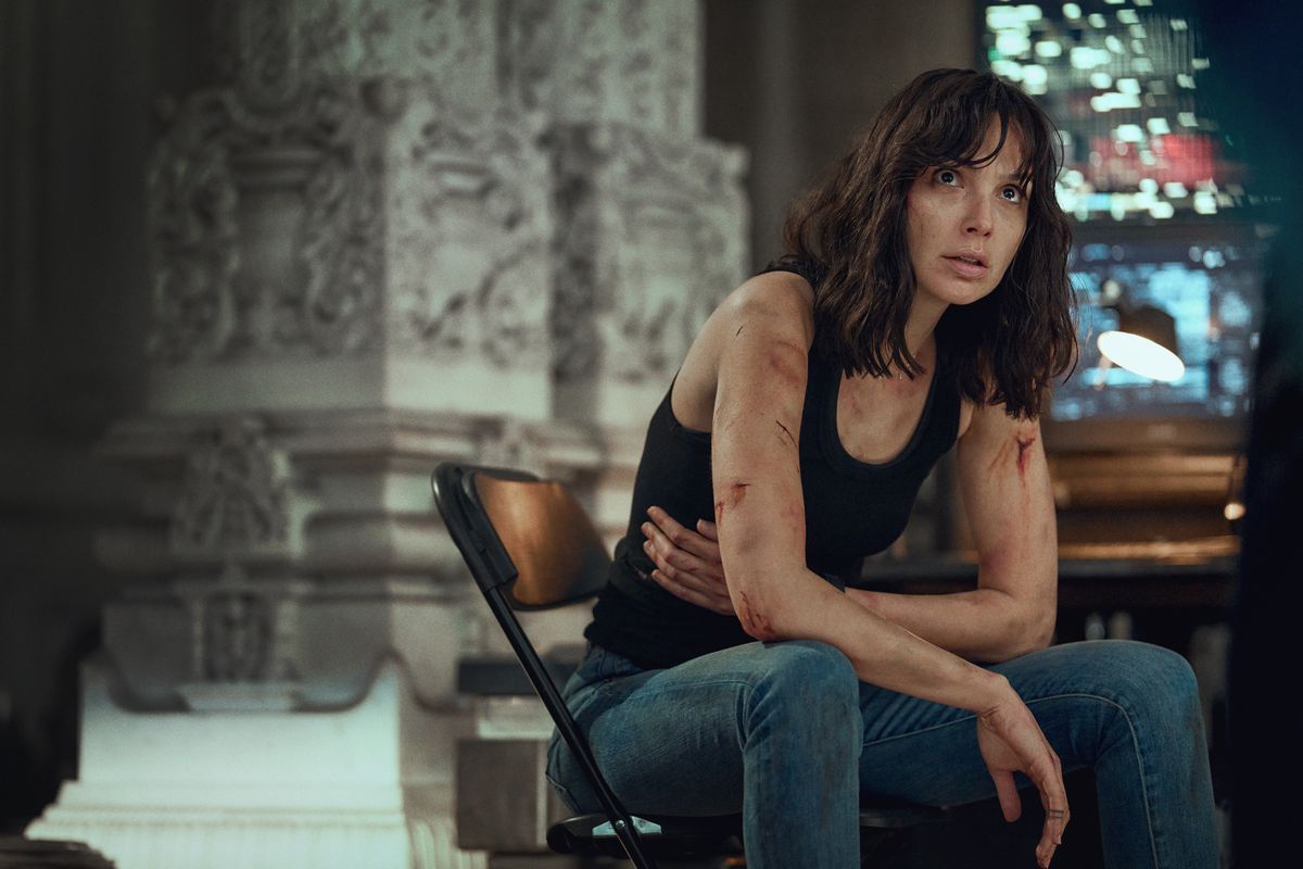 Gal Gadot, wearing a black tanktop with cuts all down her arms, holds onto her side while sitting in a chair in Heart of Stone.