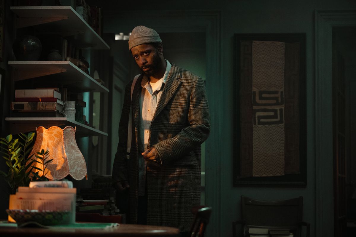 LaKeith Stanfield stands in a worn overcoat and beanie in an eerily green apartment draped in shadow in the Apple TV Plus series The Changeling