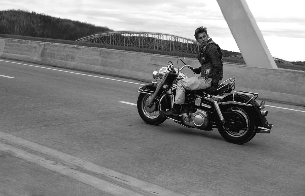 Austin Butler, in black and white, rides his Harley-Davidson across a bridge one-handed, looking back over his shoulder in a very cool way in The Bikeriders