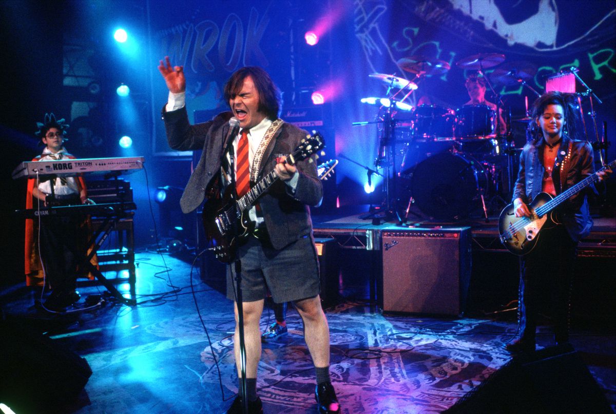 Jack Black in a suit jacket, bright red tie, and shorts, rocking out at the mic on stage in Richard Linklater’s 2003 movie School of Rock