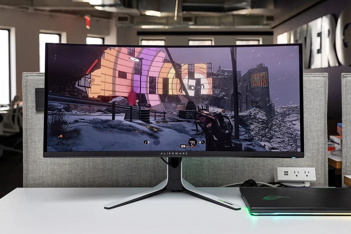 A photo of the Alienware QD-OLED AW3423DW curved gaming monitor sitting on a white desk, connected to a gaming laptop. The game Deathloop is showing on its screen.