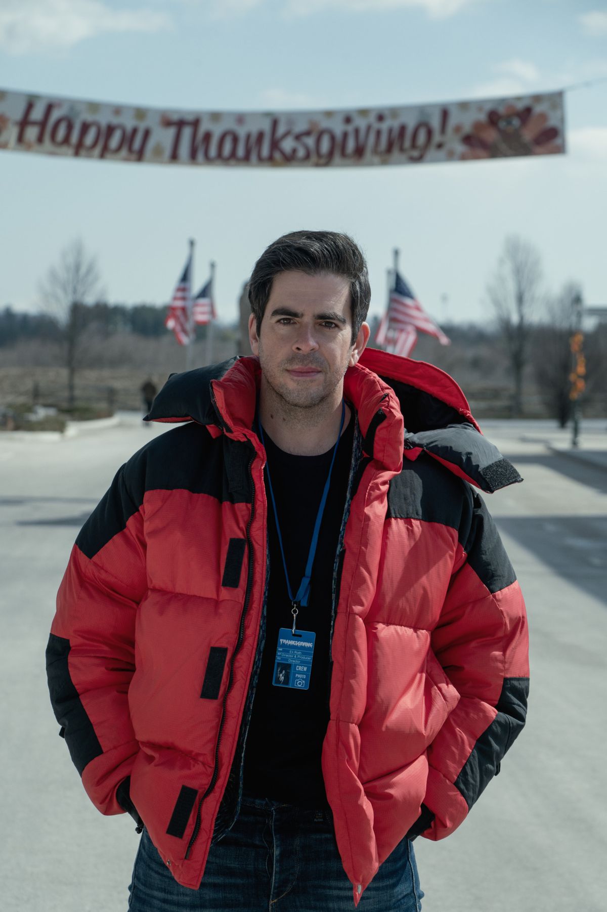 Eli Roth in a red puffy jacket on the set of Thanksgiving with a Happy Thanksgiving banner behind him
