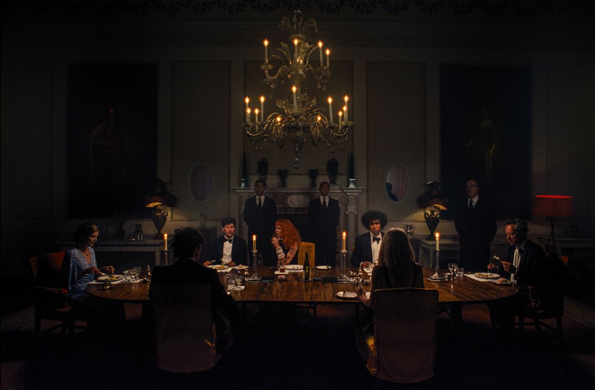 الیور (بری کیوگان) در فیلم Saltburn همراه با فلیکس (جیکوب ژاکوب الوردی) و سایر اعضای خانواده اش، همه با لباس رسمی، پشت میز ناهارخوری طویل در اتاقی بسیار تاریک نشسته است.