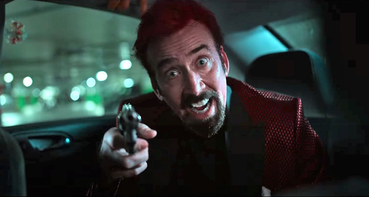 A bearded Nicholas Cage in a red suit aiming a gun from the backseat of a car and smiling maniacally in Sympathy for the Devil.