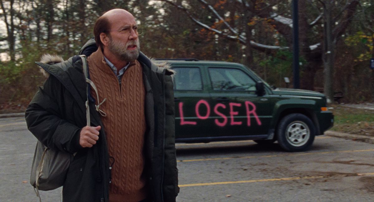 A schlubby-looking Nicolas Cage holds a backpack and stands in front of a car with “LOSER” painted on it in bright pink letters in Dream Scenario.