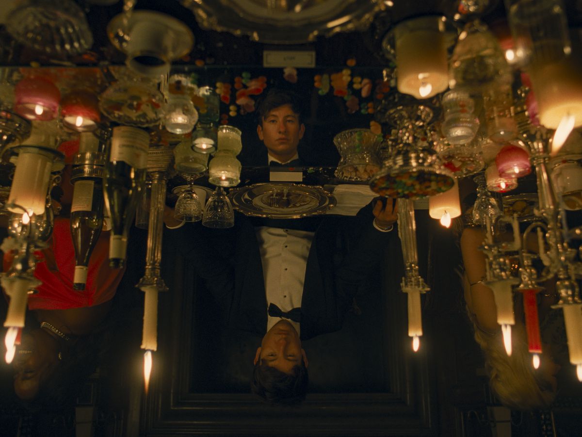 Oliver (Barry Keoghan), in black tie dress, sits at what appears to be an fancy table covered in candles of all descriptions, reflecting his face back at him —&nbsp;except the more you look, the more it’s clear that the reflection is in a different position, standing with its eyes lowered. From the movie Saltburn