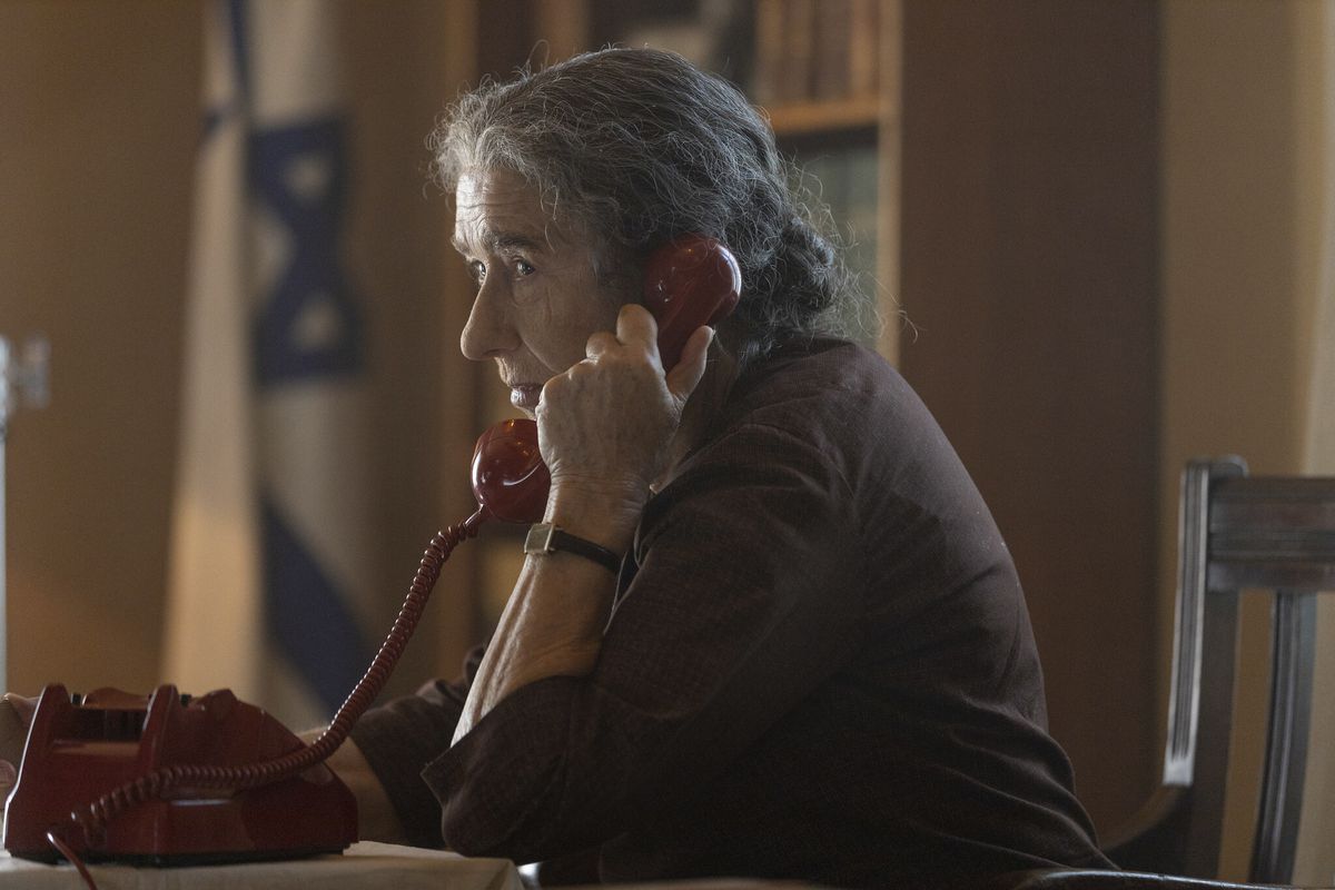 Helen Mirren as Golda Meir, sitting at a table and speaking into a red corded telephone with the flag of Israel in the background in Golda.
