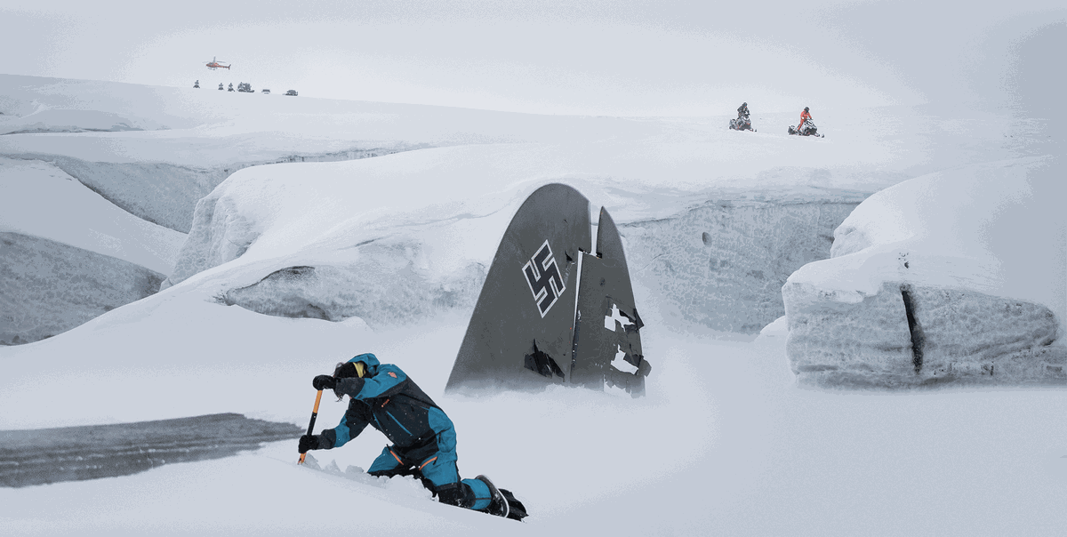 A man in a blue snowsuit shovels the wreckage of a Nazi biplane out of the snow with two figures riding snowmobiles in the distance in Operation Napoleon. 