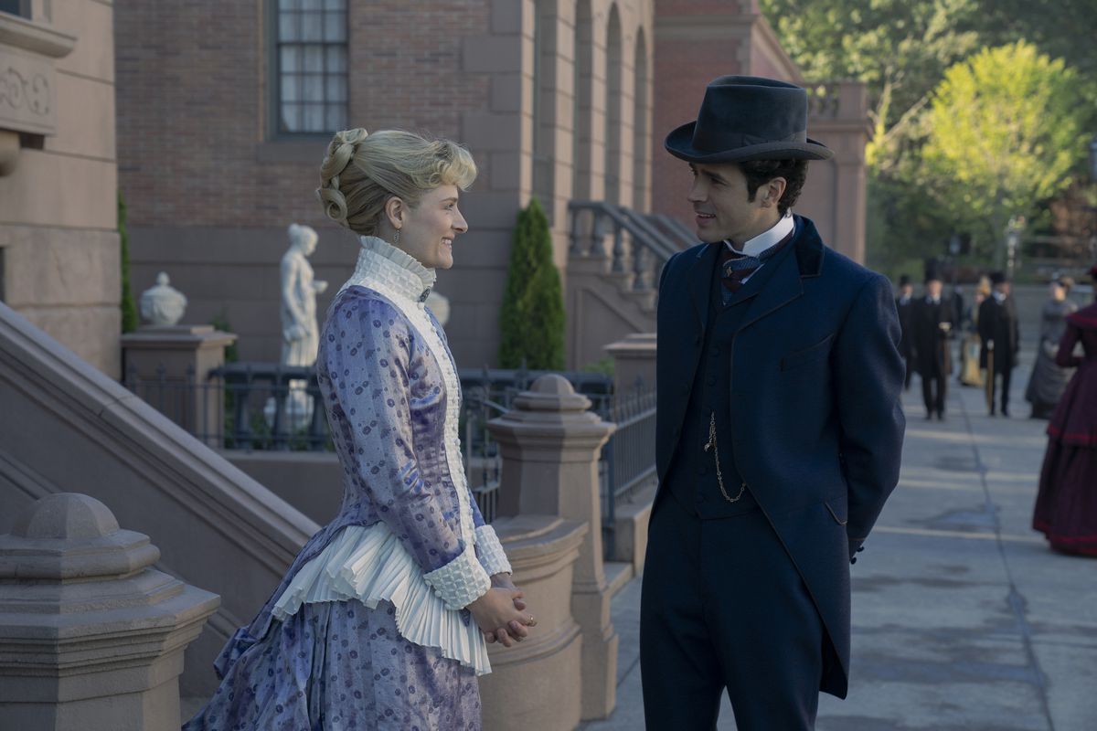Louisa Jacobson and Harry Richardson standing and smiling at each other on the street in a still from The Gilded Age season 2