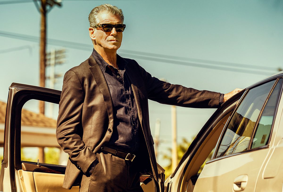 Pierce Brosnan, wearing a suit and shades, leans against a car with an open door in Fast Charlie