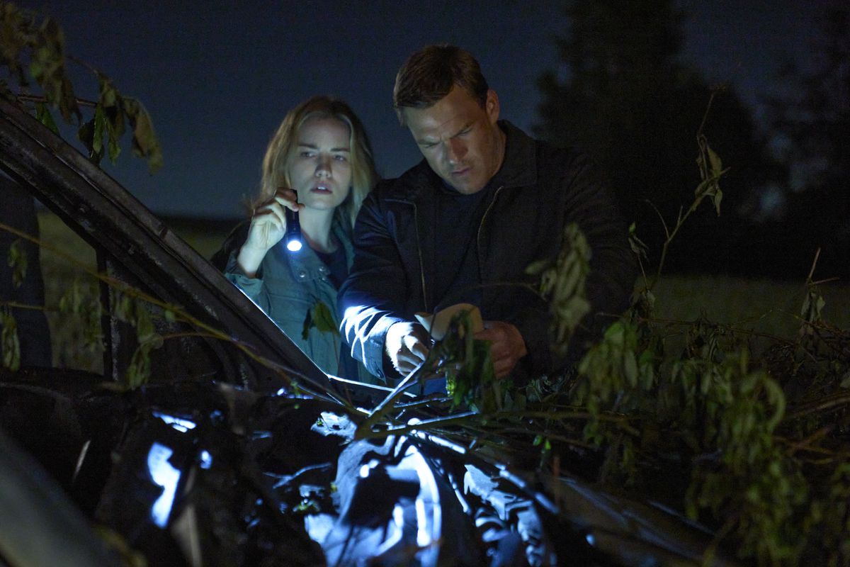 Jack Reacher, a large man wearing a collared sweater, investigates in the woods with a blonde female police officer (holding a flashlight).