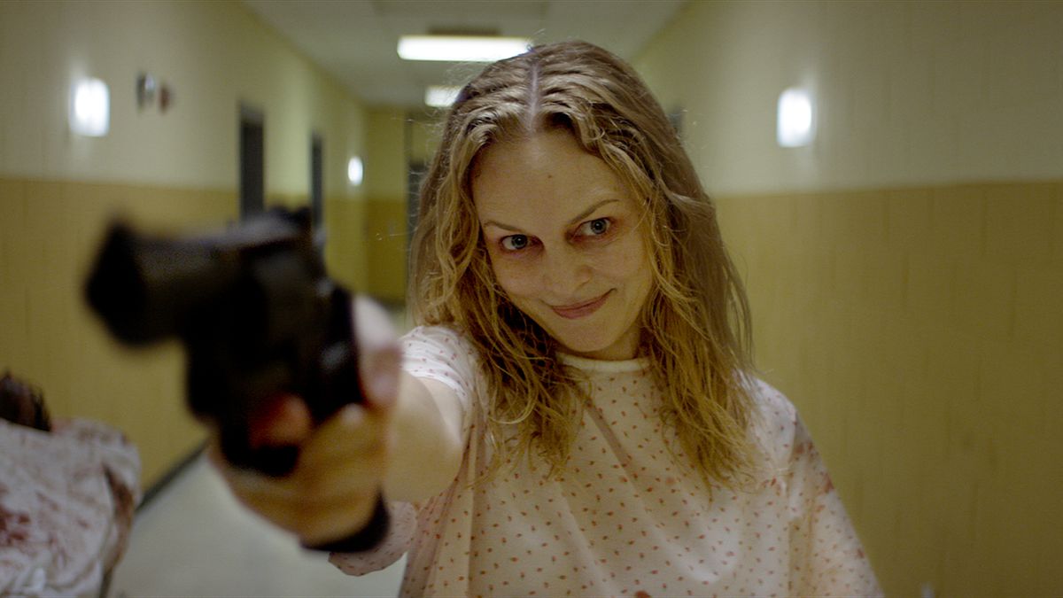 A smiling woman in a hospital gown aims a revolver in a poorly lit hallway in Suitable Flesh.