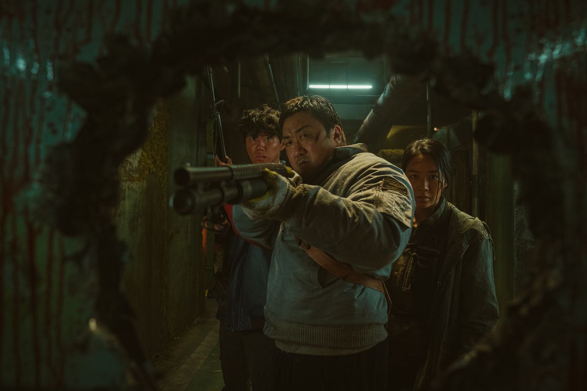 A man (Don Lee) points a shotgun through a hole while a young man and a woman stand behind him in a darkened hallway in Badland Hunters.