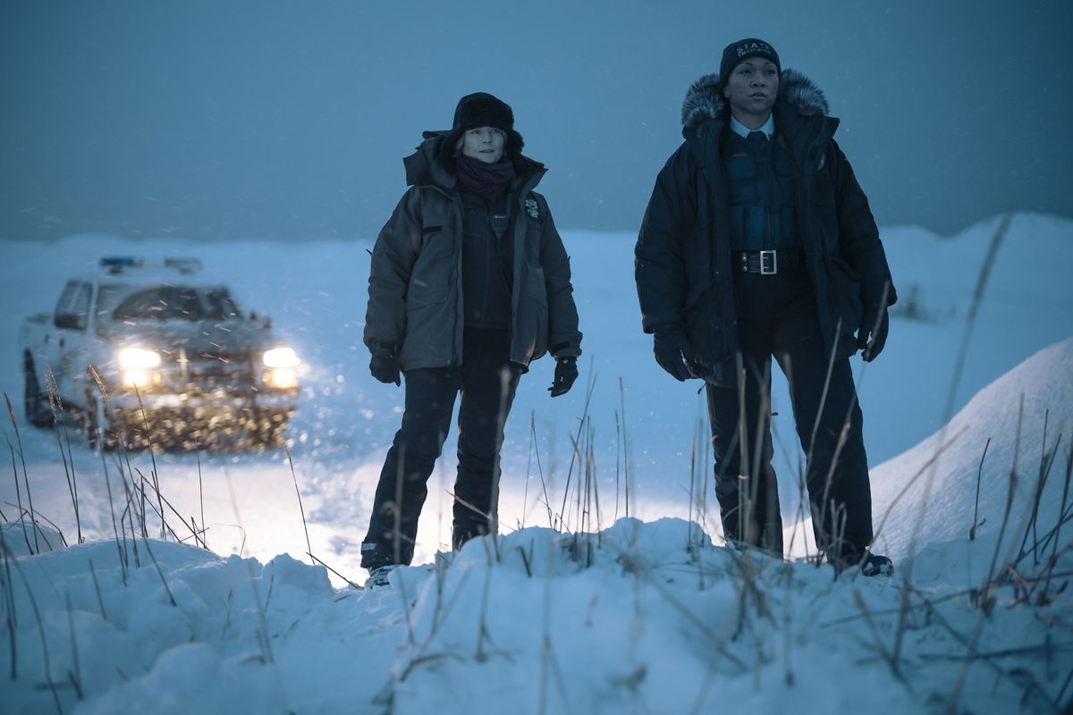 Alaska detectives Liz Danvers and Evangeline Navarro stand on a snowy hill in front of their squad truck in the HBO series True Detective: Night Country.