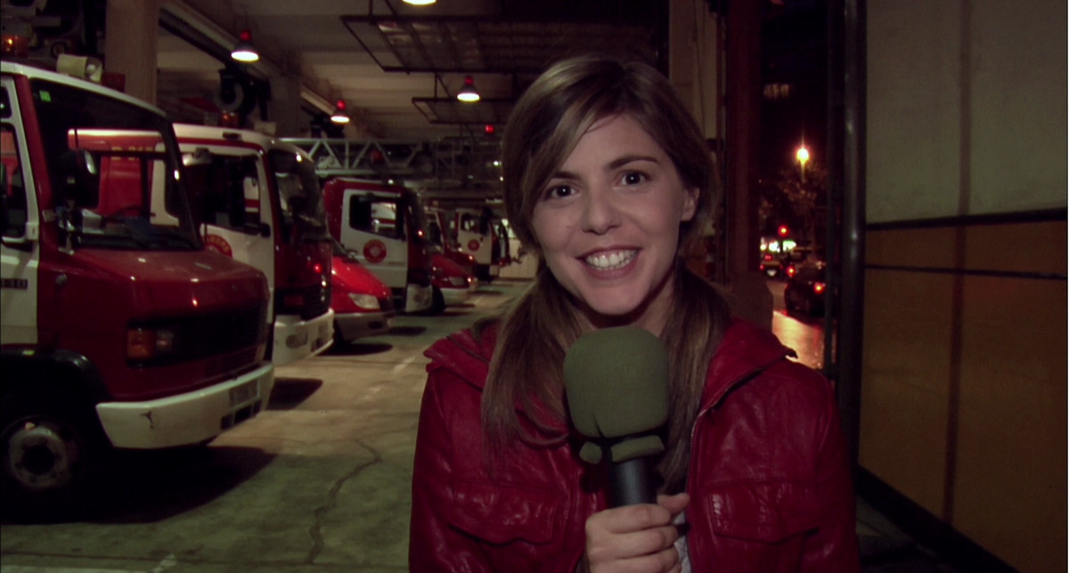زن جوانی که ژاکت قرمز بر تن دارد در یک پخش تلویزیونی از ایستگاه آتش نشانی در Rec با میکروفون صحبت می کند.