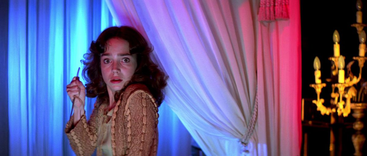 Jessica Harper holds a sharp object in her hand while looking scared in Suspiria. She stands next to a curtain, with red, blue, and white lighting around her.