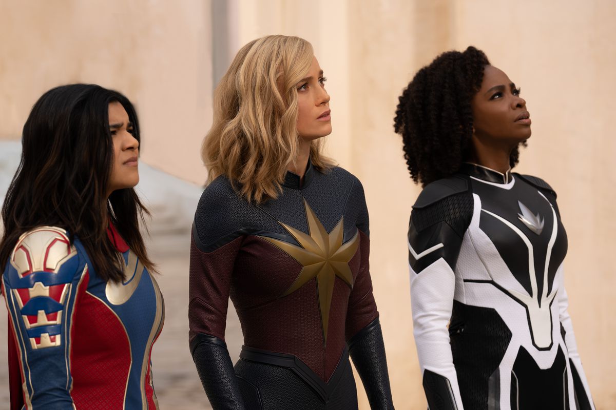 Iman Vellani as Ms. Marvel/Kamala Khan, Brie Larson as Captain Marvel/Carol Danvers, and Teyonah Parris as Captain Monica Rambeau stand together in costume, all looking up, in the Marvel Cinematic Universe movie The Marvels