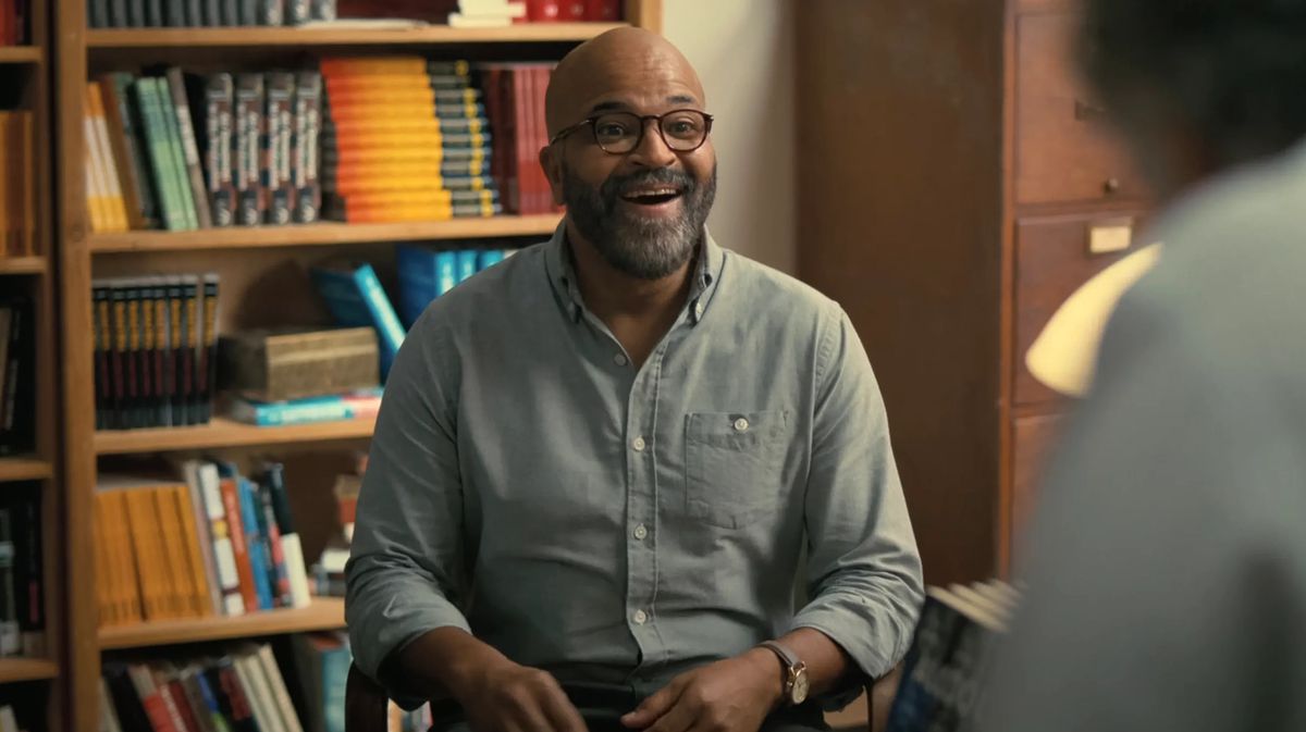 Jeffrey Wright in a blue shirt sits in his library giggling in the movie American Fiction
