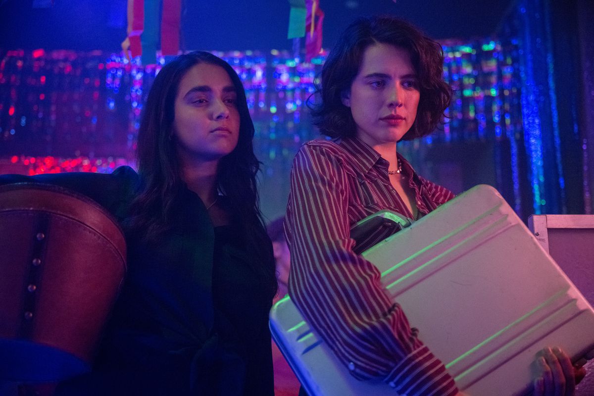 Jamie and Marian clutch a briefcase close in a bar full of bisexual lighting in a scene from Drive-Away Dolls