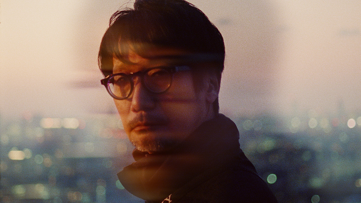 A close-up shot of a man with round glasses and short gray goatee in front of a cityscape during the afternoon.