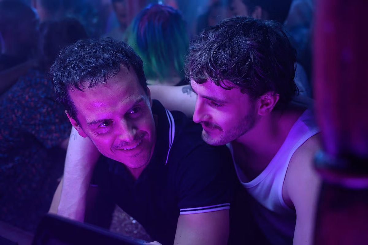 A smiling man with his arm around the shoulder of another smiling man in a nightclub filled with people.