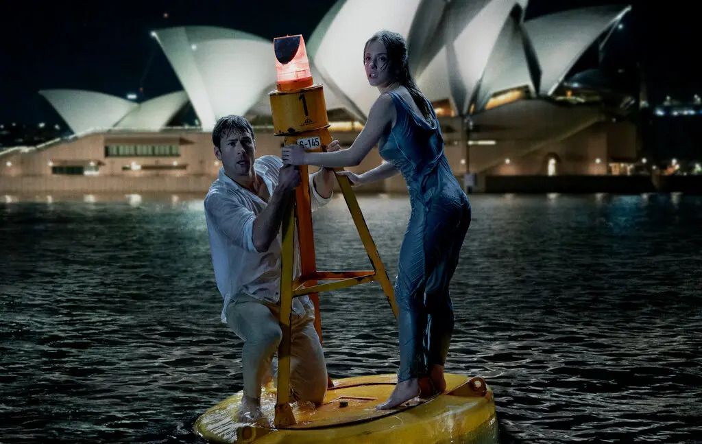 A man and a woman cling to a buoy in a bay with the Sydney Opera House visible in the background.