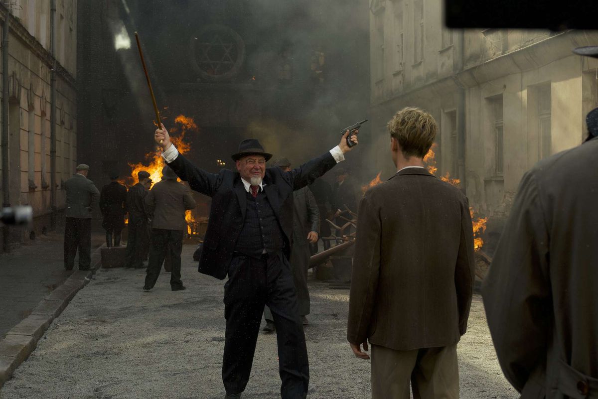 A man with a hat and a pointed white beard with no moustache raises his arms in triumph in front of a burning synagogue. He’s holding a gun