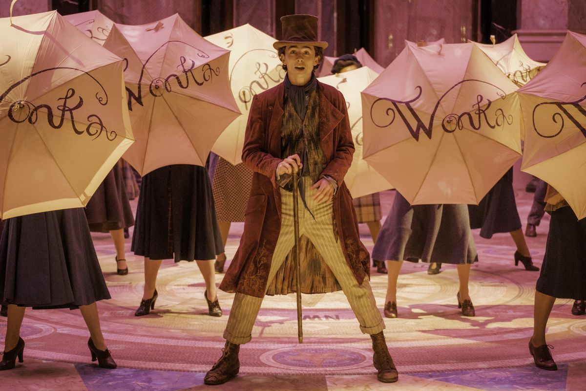 Wonka stands with his cane in front of him as dancers hold up umbrellas with ‘Wonka’ on them behind him in the movie Wonka.