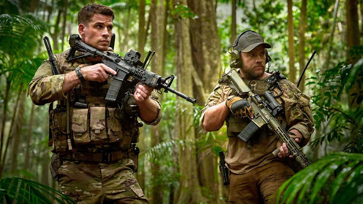 Two men in army camouflage holding rifles in a tropical forest.