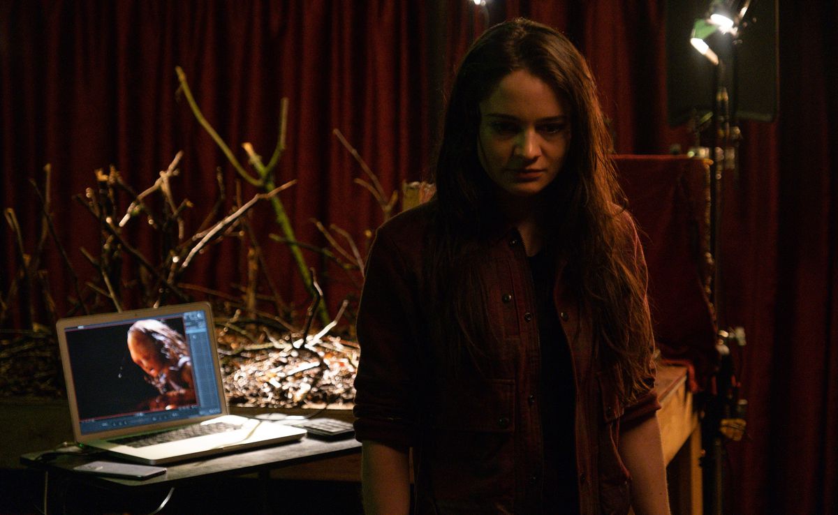 Stop-motion animator Ella (Aisling Franciosi) stands with her back to her set, a tangle of branches on a table in front of a red curtain, with a laptop open on the table, showing the warped little skull-faced girl from the movie she’s making in Stopmotion
