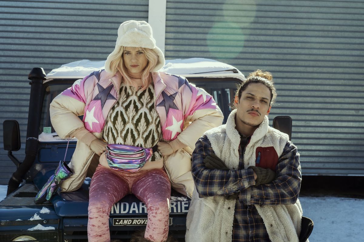 A woman with pink hair wearing a star-covered coat and a white fur hat and a striped neon fanny pack sits in the bed of a pickup truck, with a man with a top bun wearing a vest standing next to her