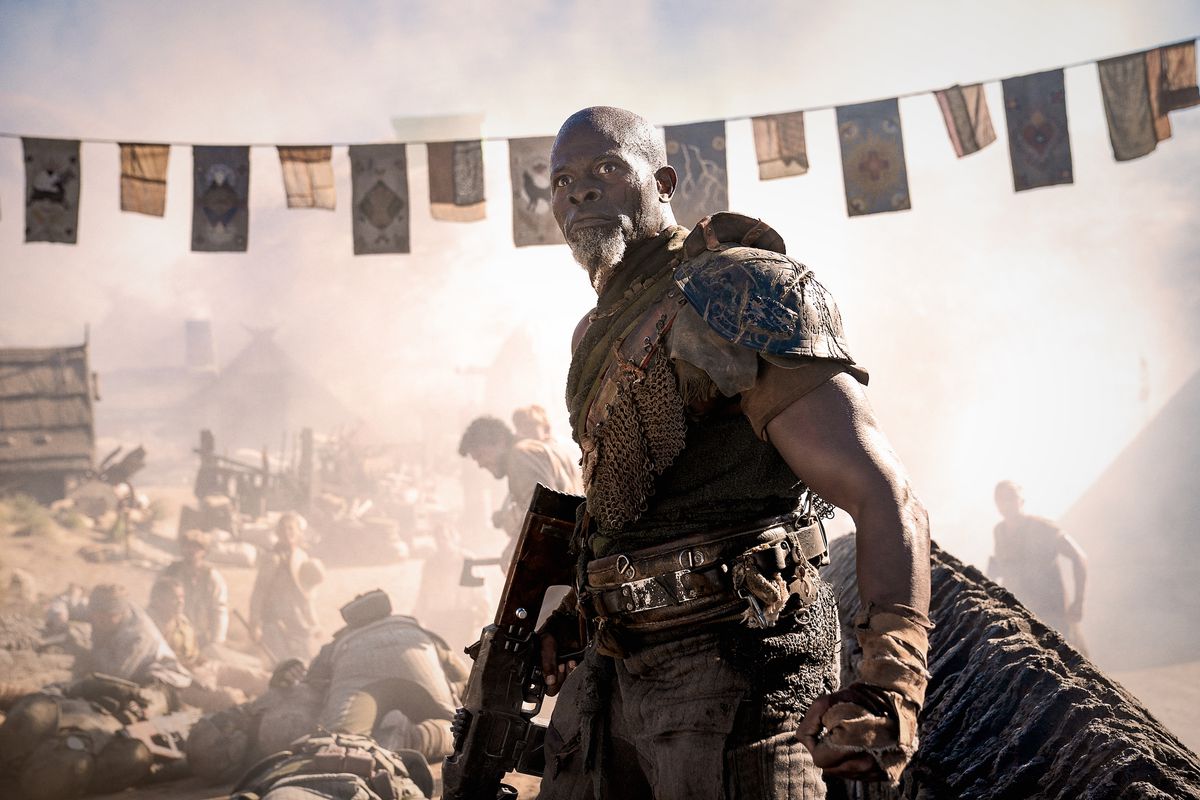 Djimon Hounsou รับบทเป็นนายพล Titus ใน Rebel Moon - ตอนที่สอง: The Scargiver ถือปืนอยู่ข้างๆ ขณะที่เขายืนอยู่ใกล้หมู่บ้านที่ถูกสังหารหมู่
